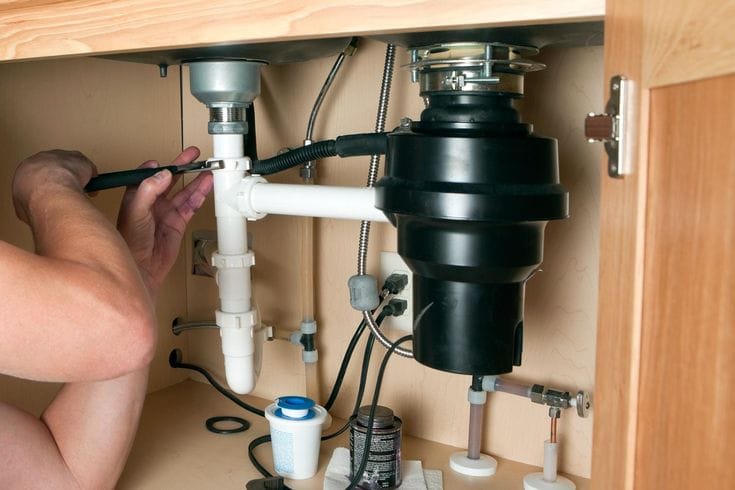 Plumber installing a garbage disposal beneath a kitchen sink in Westminster